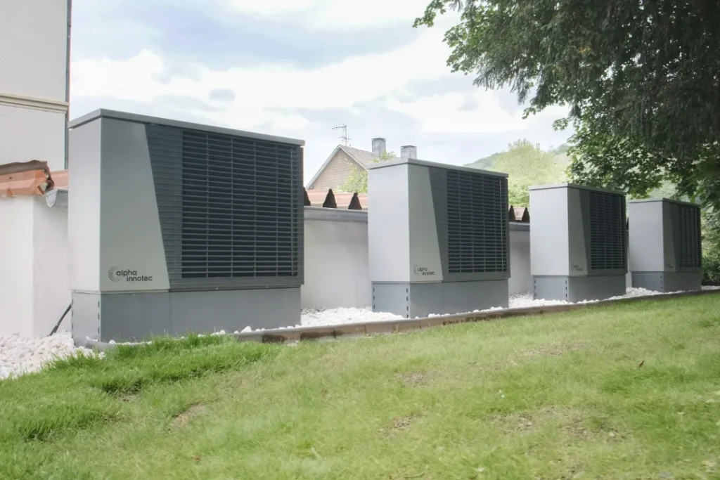 Cztery duże, szare, przemysłowe jednostki HVAC stoją wzdłuż białego budynku obok trawiastego trawnika, za nimi drzewa i dachy; scena wydaje się spokojna i użyteczna.